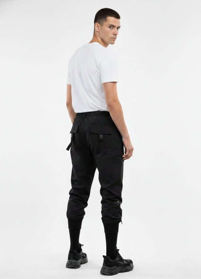 Rear view of black tactical techwear pants with buckle straps and utility pockets, styled with white t-shirt and sneakers.