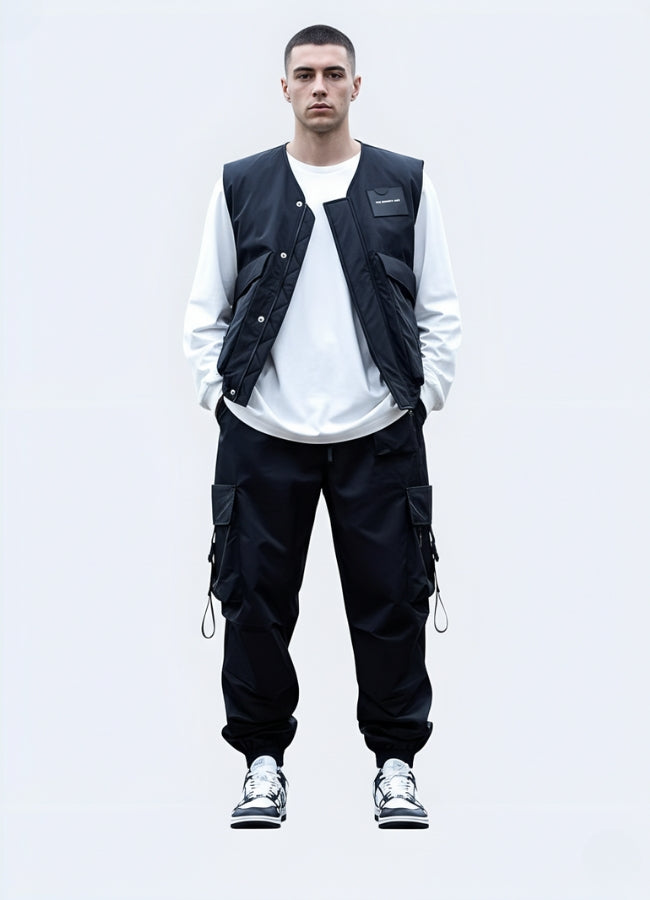 Male model in minimalist techwear outfit featuring japanese techwear pants, white long-sleeve shirt, and tactical vest, styled with urban sneakers on clean background.