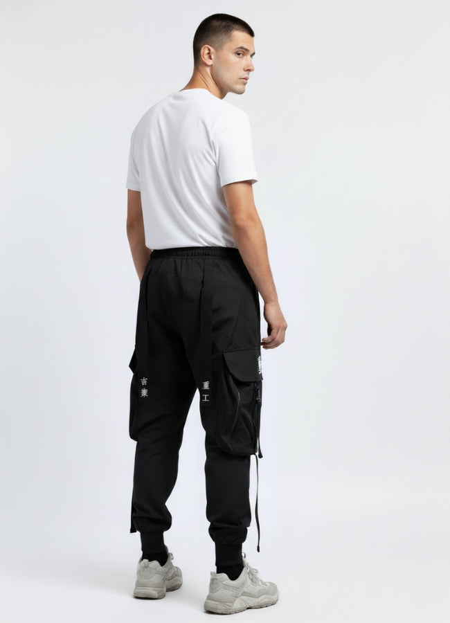 Back view of a man wearing black shinobi pants with oversized pockets, white graphic symbols, and hanging straps, paired with a white T-shirt and grey sneakers.