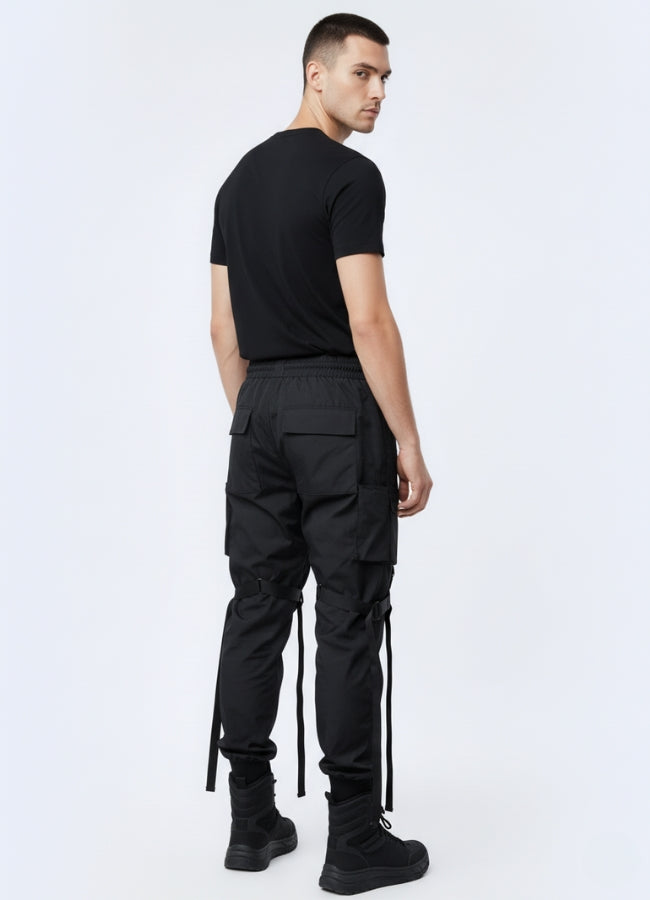 Back view of a man wearing black streetwear joggers with multiple pockets, printed patches, and adjustable strap details, paired with a black T-shirt and black tactical boots.