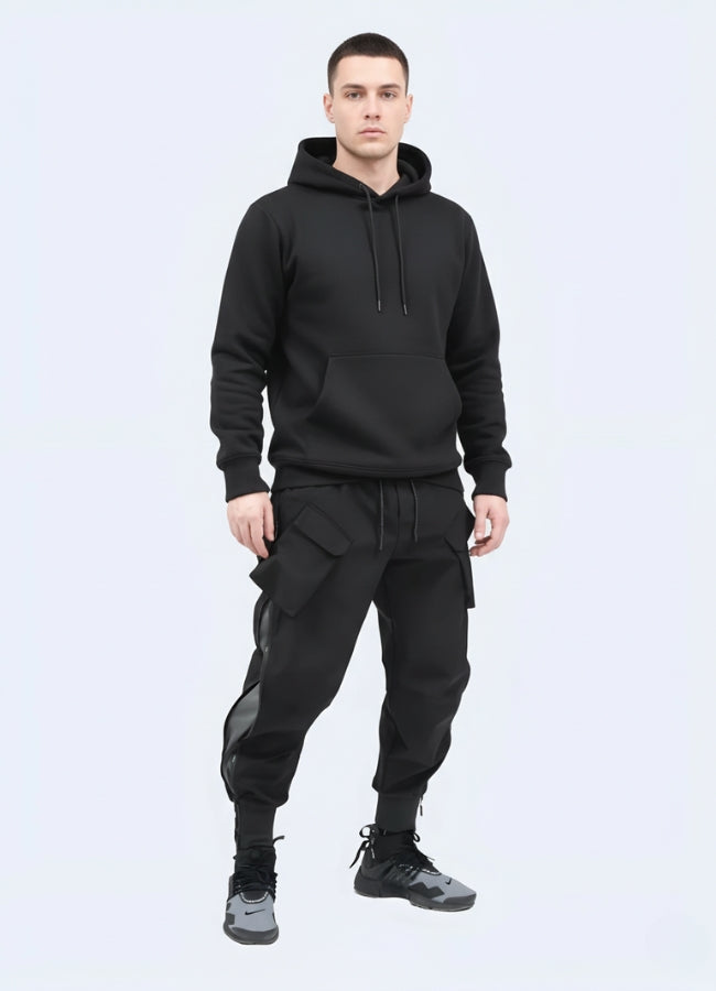 Male model wearing black techwear cargo pants with multiple pockets and tapered cuffs, styled with black hoodie and sneakers on white background.