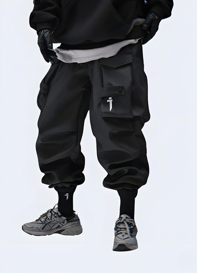A person wearing black cargo pants and black gloves, with a white undershirt peeking out, stands against a light background. The individual is wearing dark grey athletic shoes with white laces and black socks that have a white logo. The focus is on the lower half of the body, showcasing a trendy, utilitarian style.
