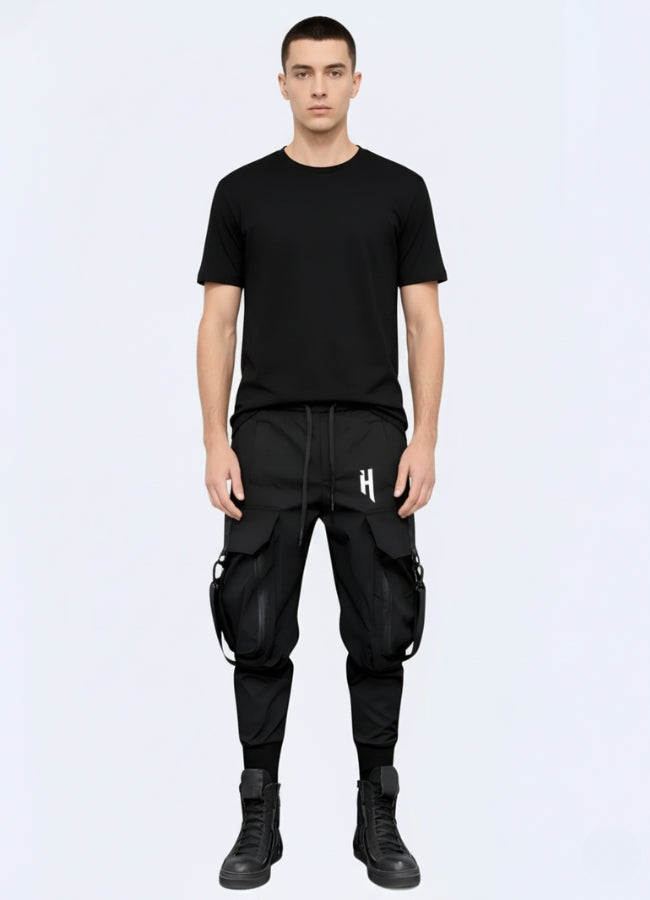 Male model wearing black techwear cargo pants with detachable straps and oversized pockets, styled with black t-shirt and high-top boots.