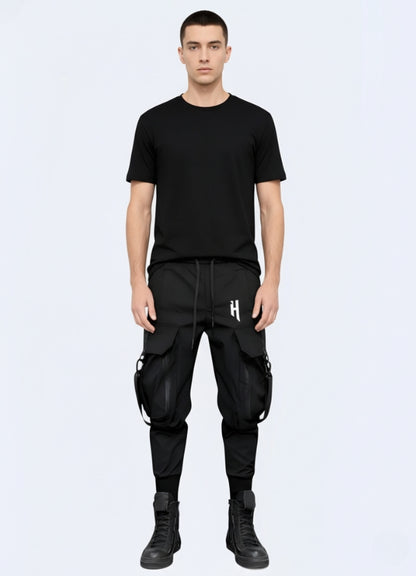Male model wearing black techwear cargo pants with detachable straps and oversized pockets, styled with black t-shirt and high-top boots.