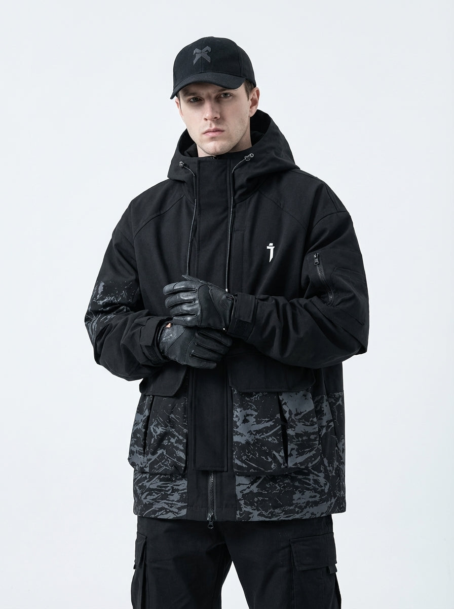 Front view of male model wearing a black techwear jacket with grey abstract camouflage pattern on the lower half and sleeves, featuring multiple cargo pockets, a white logo on the chest, black cargo pants, and tactical gloves.