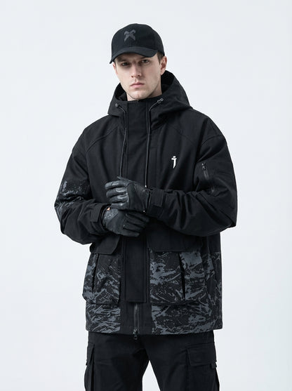 Front view of male model wearing a black techwear jacket with grey abstract camouflage pattern on the lower half and sleeves, featuring multiple cargo pockets, a white logo on the chest, black cargo pants, and tactical gloves.