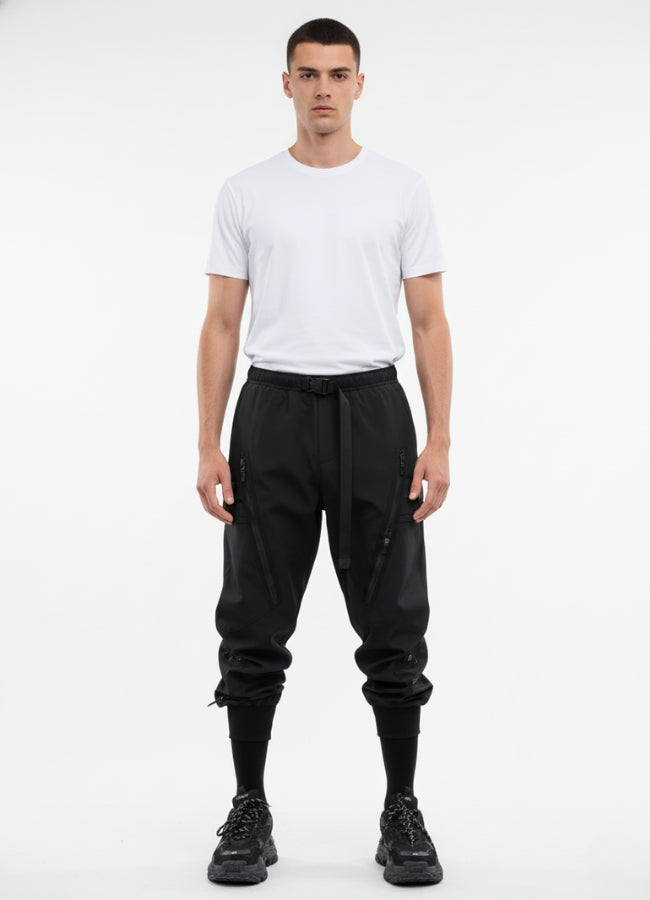 Male model wearing black techwear cargo pants with front zippers and belted waistband, paired with white t-shirt and black sneakers.