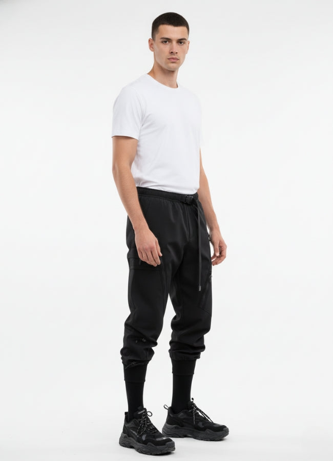 Side angle of black techwear pants featuring adjustable belt, multi-pocket design, and tapered ankle cuffs.