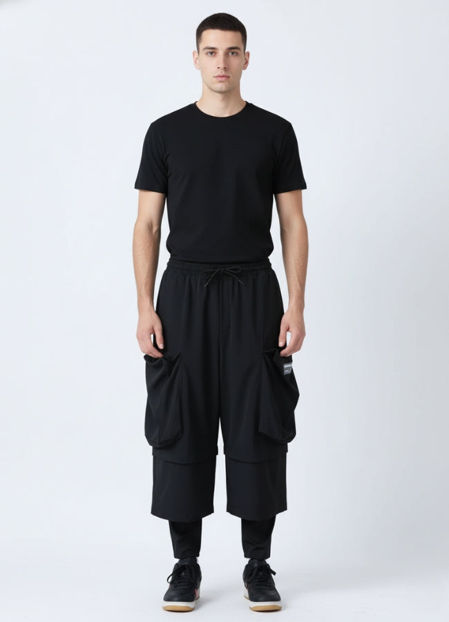 Front view of a man wearing black loose techwear pants with oversized cargo pockets, paired with a black T-shirt and black sneakers