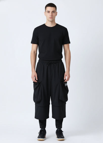 Front view of a man wearing black loose techwear pants with oversized cargo pockets, paired with a black T-shirt and black sneakers