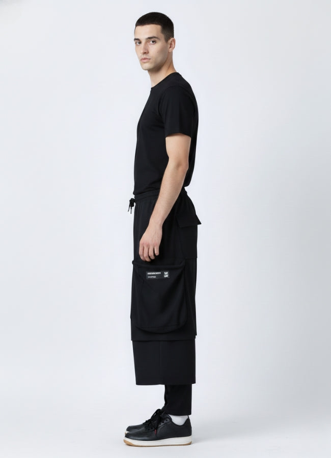 Side view of a man wearing black loose techwear pants featuring large side cargo pockets, styled with a black T-shirt and black sneakers.