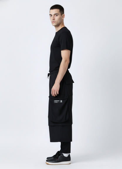 Side view of a man wearing black loose techwear pants featuring large side cargo pockets, styled with a black T-shirt and black sneakers.