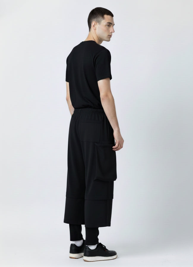 Back view of a man wearing black loose techwear pants with a rear pocket and wide-leg design, shown with a black T-shirt and black sneakers.