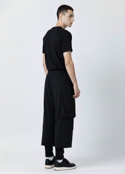 Back view of a man wearing black loose techwear pants with a rear pocket and wide-leg design, shown with a black T-shirt and black sneakers.