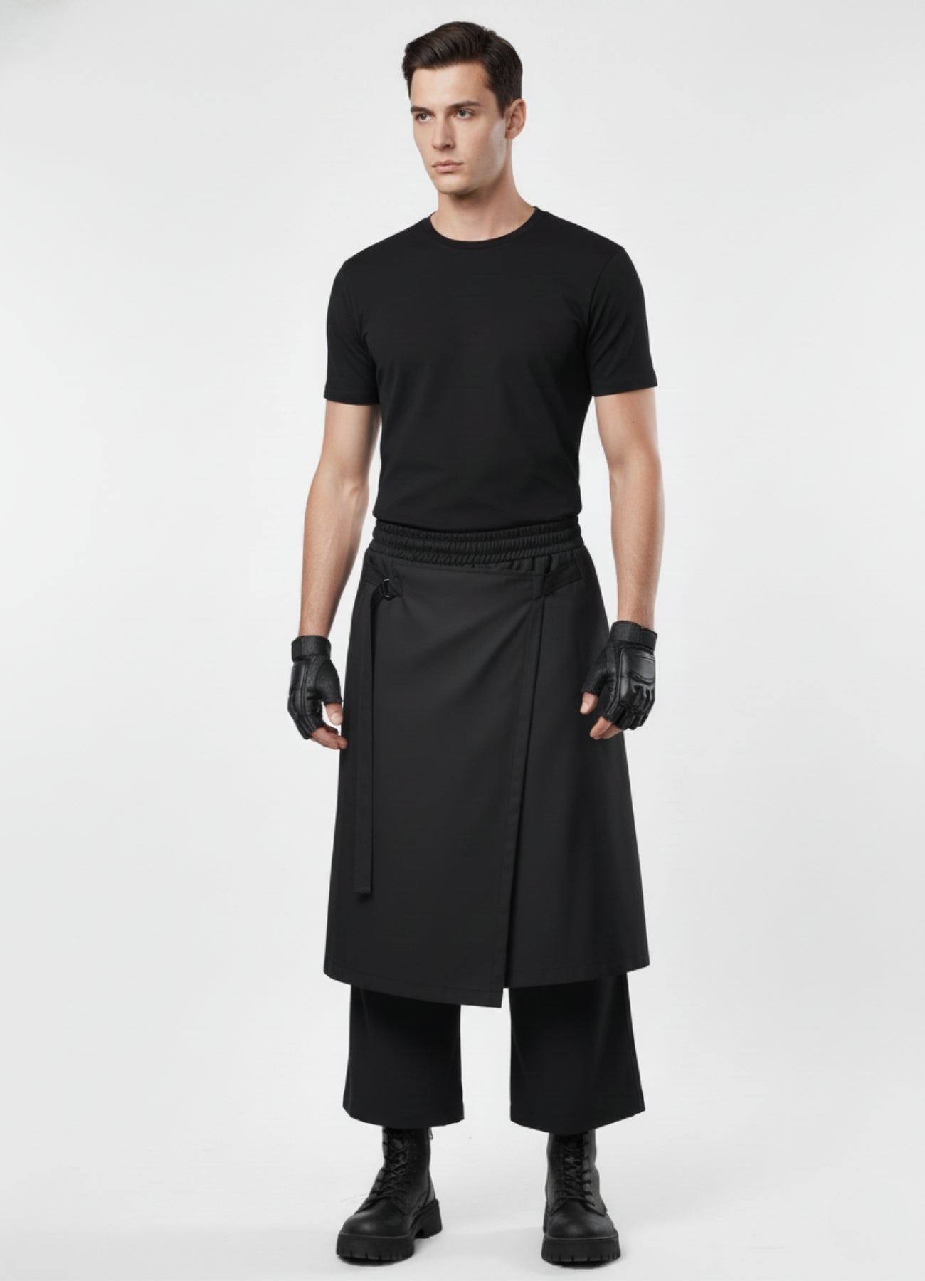 Male model wearing black wrap techwear skirt over wide-leg pants, styled with fitted black tee, gloves, and combat boots.