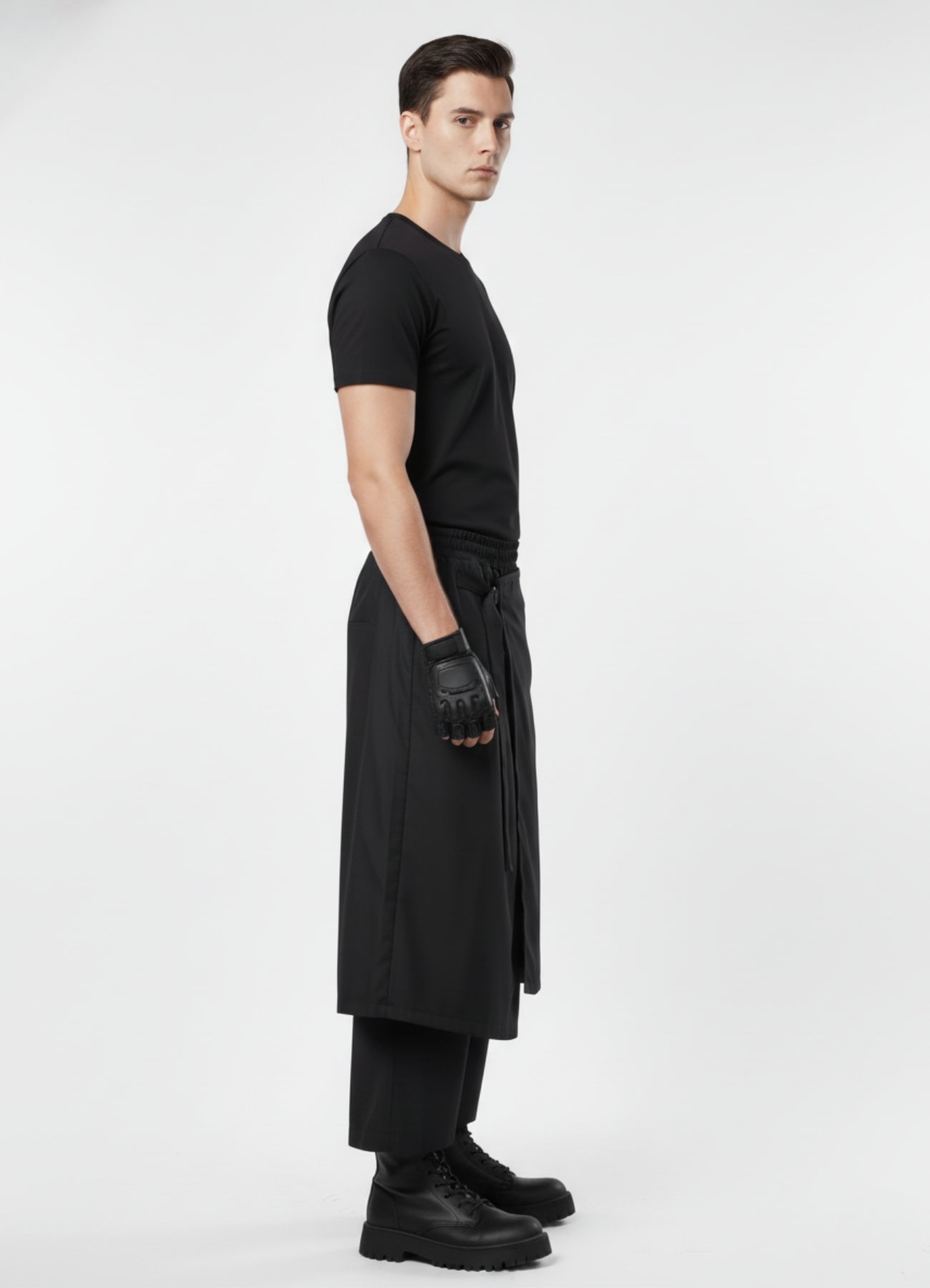 Side angle of black layered techwear skirt pants with tie-belt detail, paired with black t-shirt, gloves, and chunky boots.