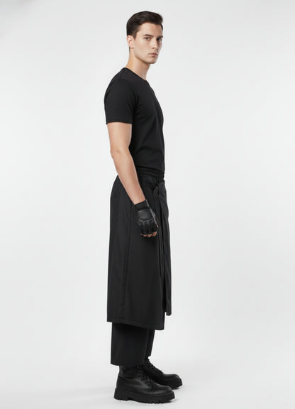 Side angle of black layered techwear skirt pants with tie-belt detail, paired with black t-shirt, gloves, and chunky boots.