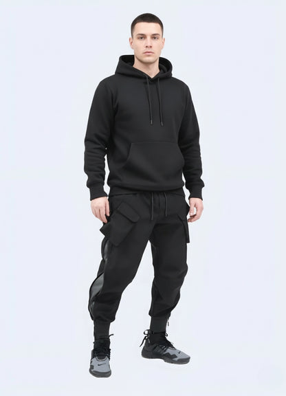 Male model wearing black techwear cargo pants with multiple pockets and tapered cuffs, styled with black hoodie and sneakers on white background.