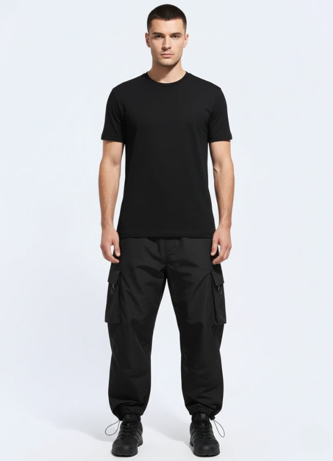 Male model wearing black techwear urban cargo pants with oversized flap pockets and elastic cuffs, styled with minimalist black tee and sneakers.