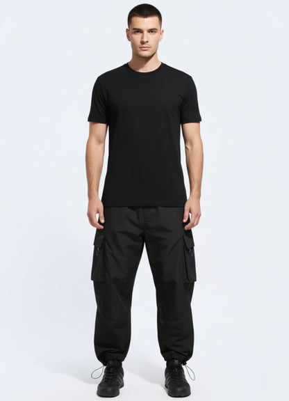 Male model wearing black techwear urban cargo pants with oversized flap pockets and elastic cuffs, styled with minimalist black tee and sneakers.