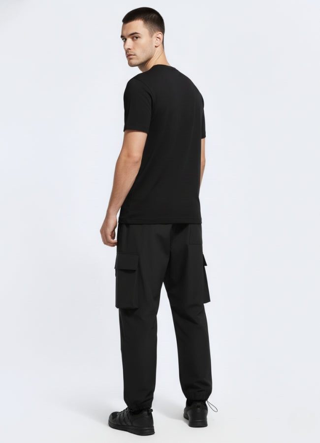 Rear view of black techwear urban cargo pants highlighting adjustable cuffs and dual pocket design, styled with black tee and shoes.