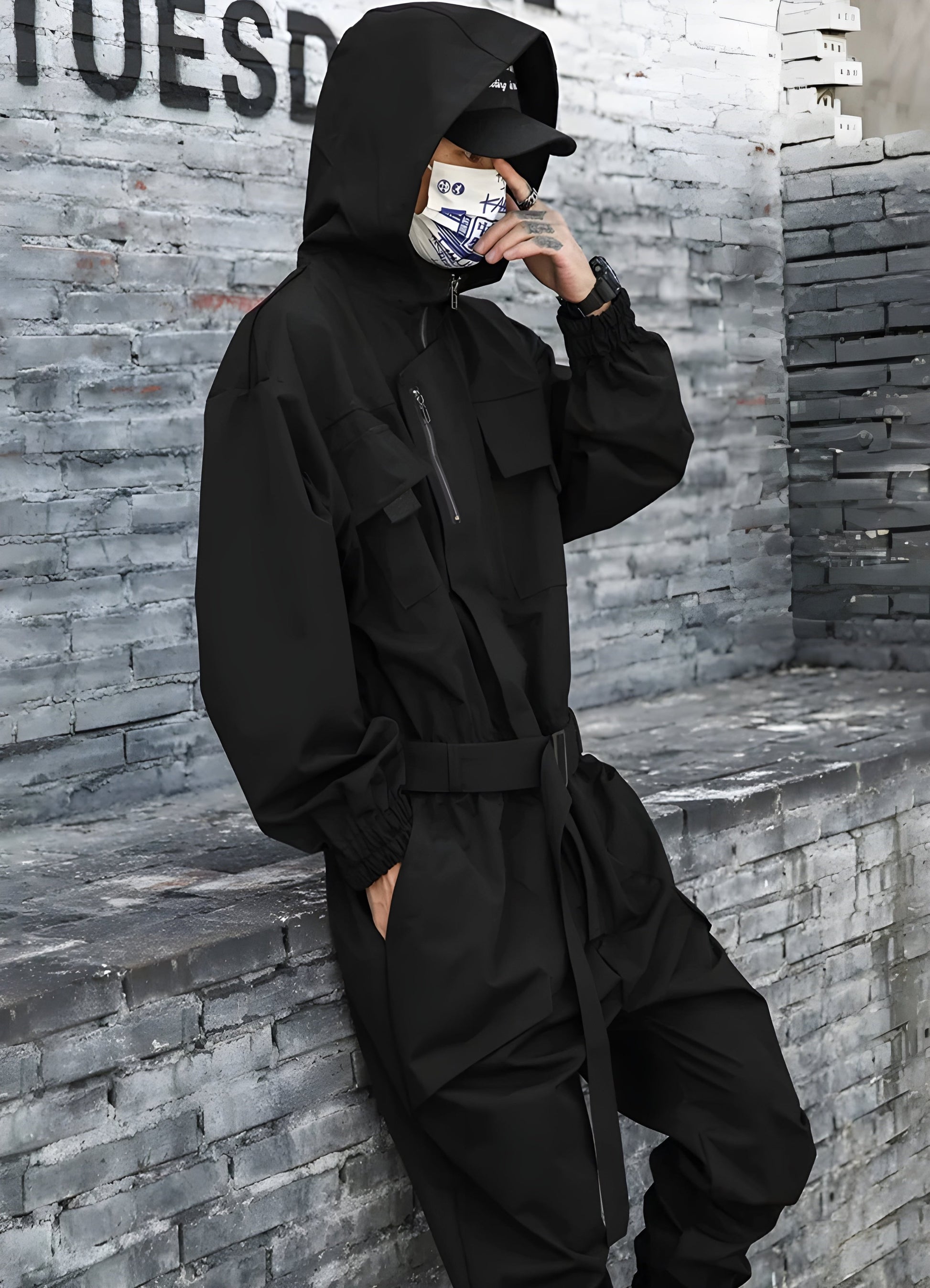 The coverall features a relaxed, oversized fit with multiple large pockets, adjustable straps, and a mix of urban design elements.